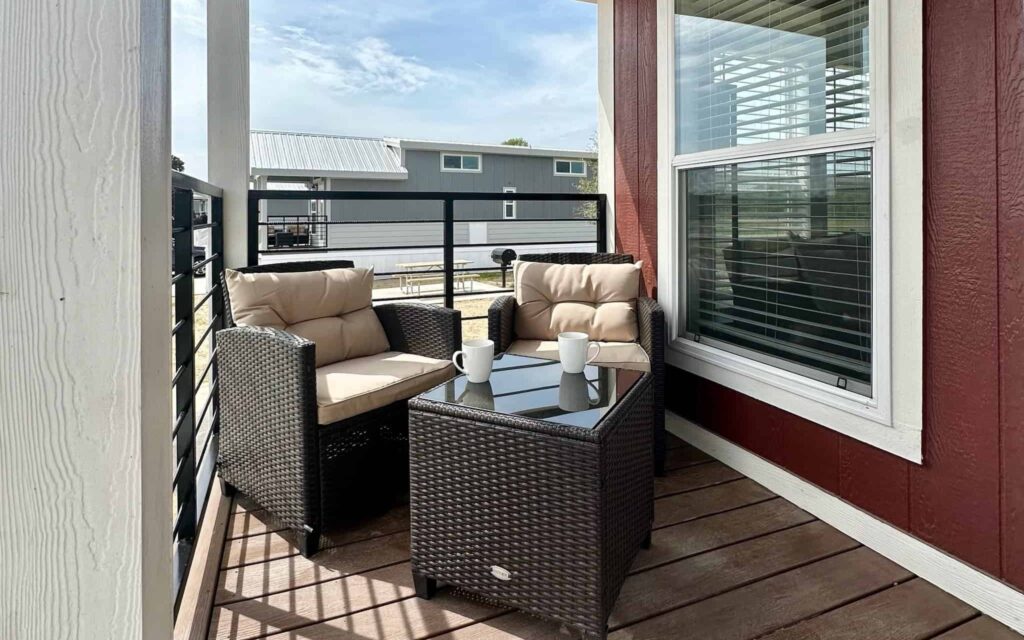 A cozy porch with two wicker chairs and a glass-top table holding coffee mugs. It's adjacent to a red building with large windows. A modern building is visible in the background under a clear sky.