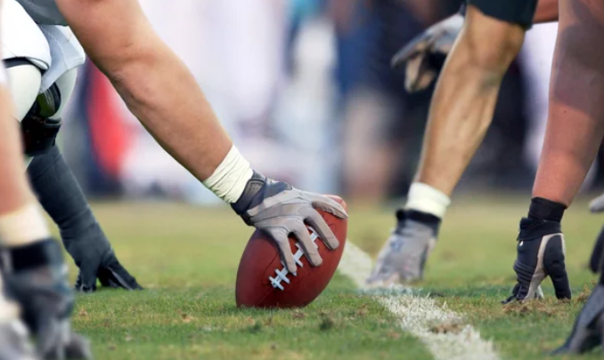 Football players on the field in a crouched position, hands on the ground and ready for the snap. A close-up of the football on the line of scrimmage, surrounded by players wearing gloves and cleats on grassy turf.