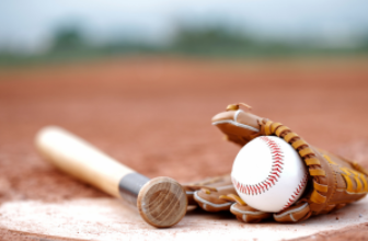 A baseball bat, glove, and baseball rest on the dirt near home plate on a baseball field, with a blurry background of grass and sky.